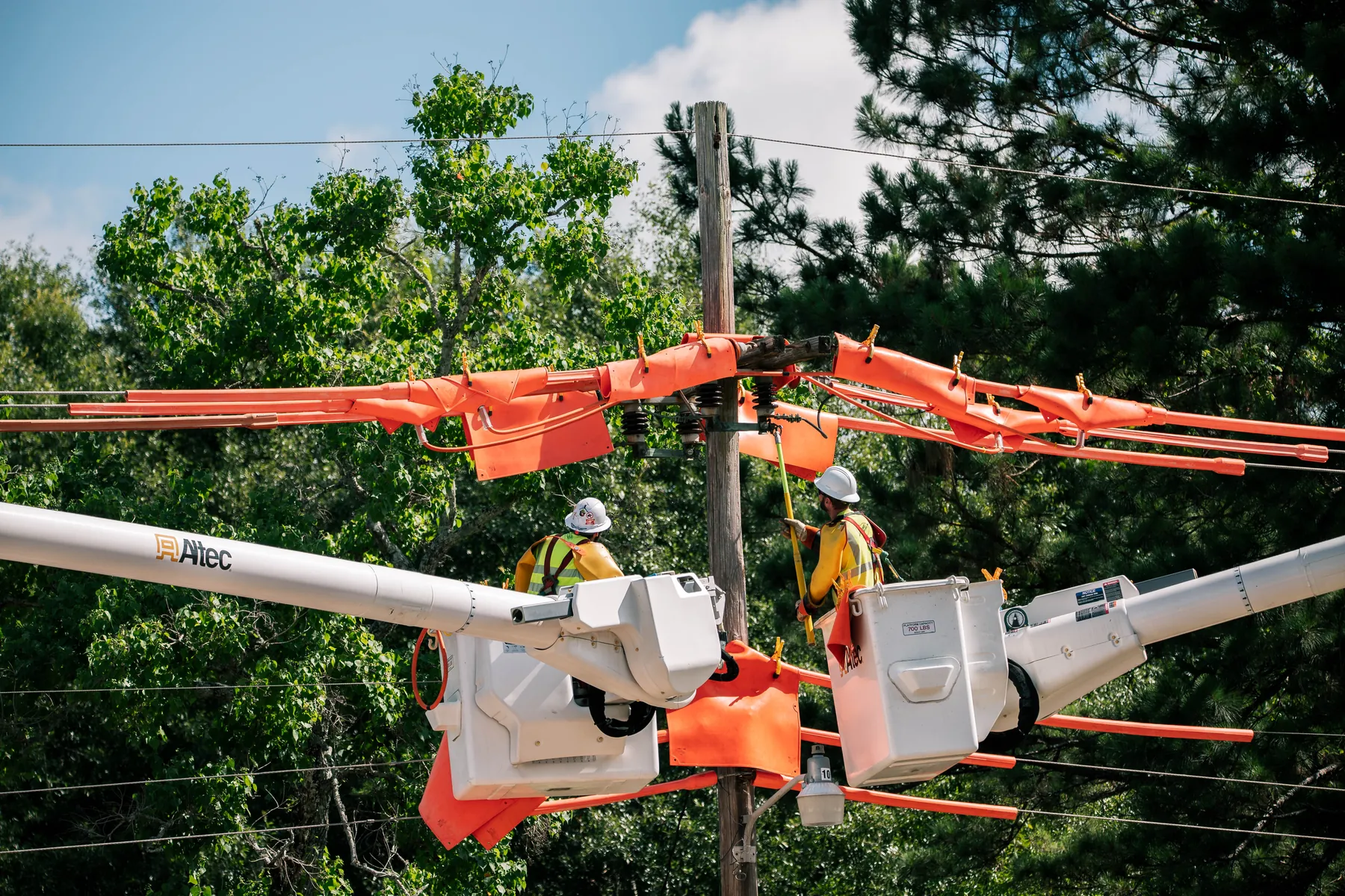 Two lineworkers in bucket trucks working on a distribution pole fitted with orange protective line covers