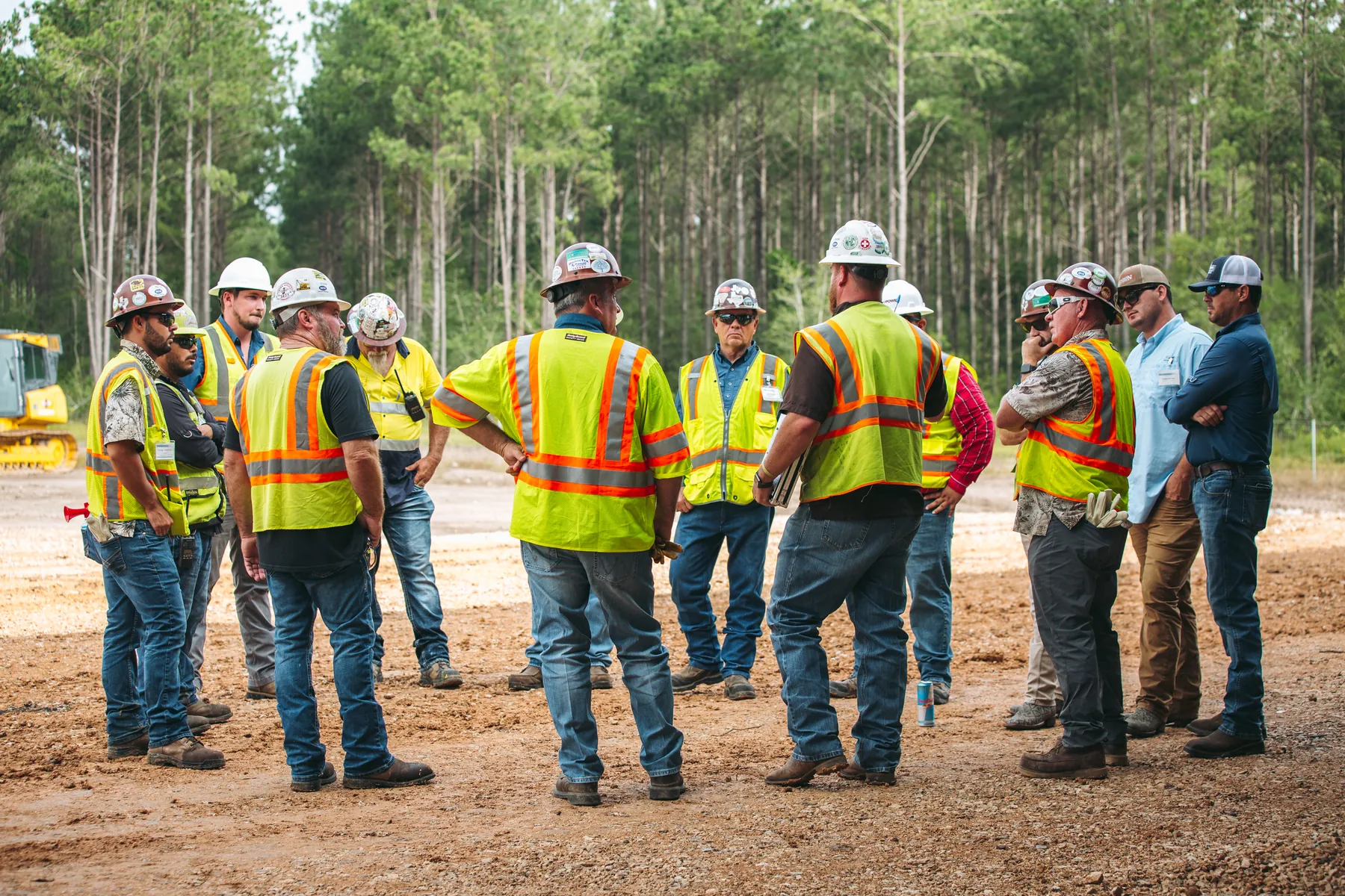 A utility crew in hard hats and safety vests standing in a circle for a job briefing on a gravel job site